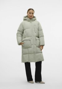 Světlé zelené péřové sako s kapucí, zipovým zapínáním a velkými předními kapsami. Obsahuje prošívaný design a oversize střih.