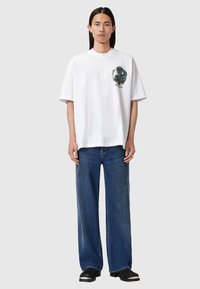 Tricou alb din bumbac cu croială supradimensionată și mâneci scurte, având un design grafic verde pe piept; purtat cu blugi denim largi de culoare albastră.