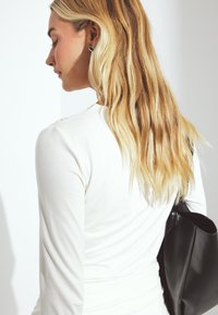 White long-sleeve top with a ruched design, paired with a black handbag. Hair is wavy and shoulder-length, against a light background.