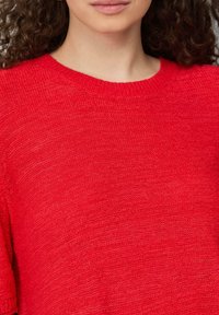 Czerwony sweter dzianinowy z krótkimi rękawami, z okrągłym dekoltem i lekko teksturowaną powierzchnią. Brak widocznych wzorów lub akcentów.