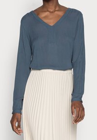 Blouse bleue à manches longues avec un col en V et un tissu texturé. Portée avec une jupe plissée crème, mettant en valeur un design de tenue à couches.