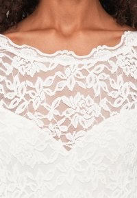 Bela čipkasta bluza z rožnatimi vzorci, valovitim izrezom in zapletenimi podrobnostmi, ki ima prozoren videz in plastev oblikovanja.