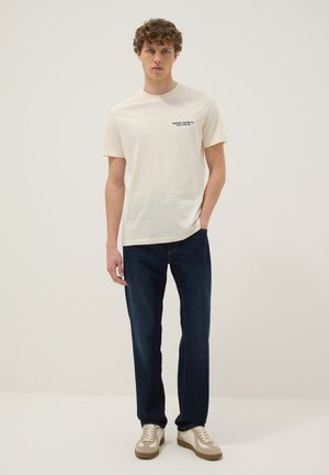 Joven de pie vestido con una camiseta blanca de manga corta con texto en el pecho, vaqueros azul oscuro y zapatillas blancas sobre fondo liso.