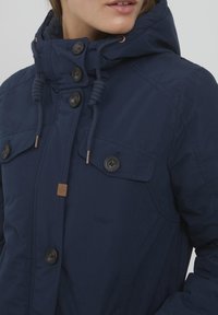 Tamsiai mėlyna parka su kapišonu, su priekiniais mygtukais, dviem krūtinės kišenėmis ir raišteliais. Pagaminta iš lygaus, izoliuoto audinio.