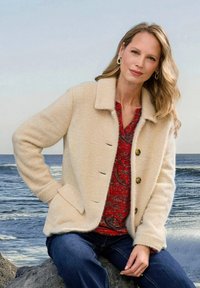 Beige wollen blend jasje met een kraag, drie knopen en twee zakken, gedragen over een rode gemusterde top en blauwe jeans. Oceaan op de achtergrond.