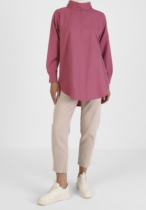 Personne portant un haut tunique rose pâle à manches longues, un pantalon beige raccourci et des baskets blanches sur un fond clair uni.