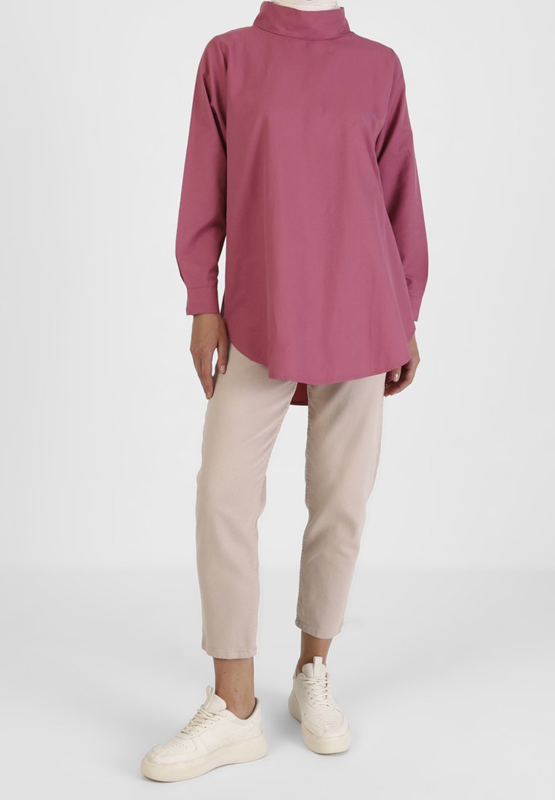 Personne portant un haut tunique rose pâle à manches longues, un pantalon beige raccourci et des baskets blanches sur un fond clair uni.