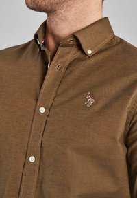 Brown cotton button-up shirt with a pointed collar, featuring a small embroidered logo on the chest and subtle contrasting stitching.