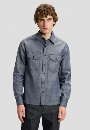 Giovane uomo con capelli ricci che indossa una camicia blu con bottoni e maniche lunghe e pantaloni scuri, in piedi contro uno sfondo chiaro e semplice.