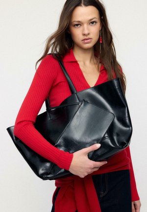 Giovane donna con top rosso a coste che tiene una grande borsa tote in pelle nera e una pochette abbinata su sfondo chiaro.