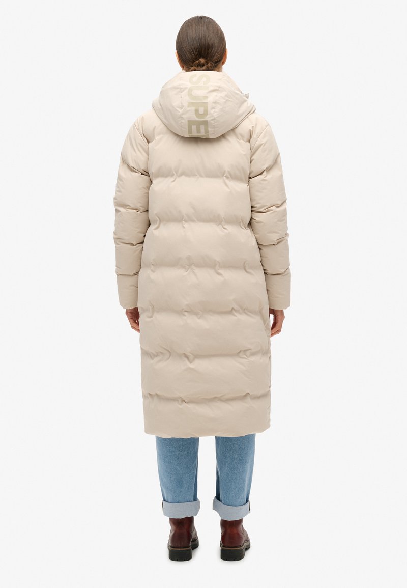 Beżowa kurtka puchowa z kapturem, z logo "SUPER" na plecach, pikowany design i długi krój, w połączeniu z podwiniętymi dżinsami i botkami.