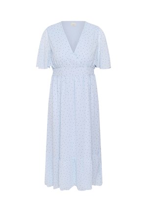 Femme aux cheveux tirés en arrière portant une robe blanche à pois noirs avec de courtes manches volantées sur un fond clair uni.