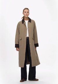 Manteau long beige avec un col marron foncé, doté de boutons pression et de deux poches avant. Le mannequin porte un haut clair et un jean foncé.