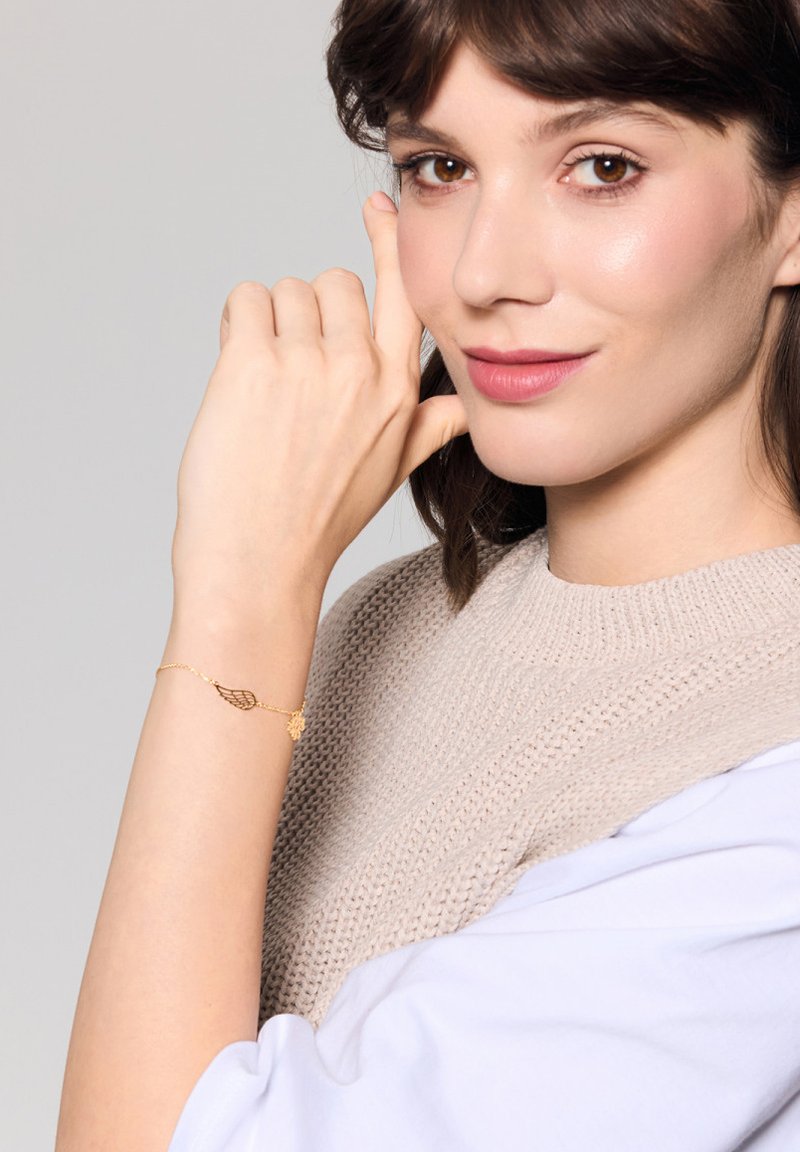 Złota bransoletka o delikatnym wzorze skrzydła i akcentem kwiatowym, noszona na jasnej skórze. Sweter w miękkim beżu na białym tle.