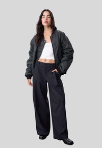 Veste bomber noire sur un crop top blanc, associée à un pantalon large à rayures marines et à des bottines noires. Fond gris neutre.