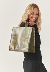 Metallic olivgrüne Tragetasche mit strukturierter Form, glatter Textur und doppelten Griffen, mit dezentem Branding auf der Vorderseite.