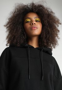 Felpa nera con cappuccio e vestibilità rilassata, con un pannello frontale testurizzato e dettaglio con coulisse. Il modello mostra una folta chioma riccia.