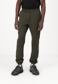 Olive green sweatpants made of soft fabric, featuring a drawstring waist and tapered cuffs. Paired with black athletic shoes.