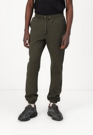 Olijfgroene sweatpants van zacht materiaal, met een trekkoord in de taille en taps toelopende boorden. Gepaarde met zwarte sportschoenen.