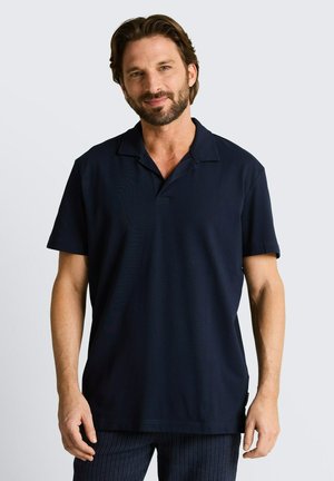 Mężczyzna z brodą w ciemnogranatowej koszulce polo z krótkim rękawem stojący na jednolitym, jasnoszarym tle.