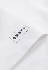 Tissu blanc avec une étiquette rectangulaire cousue, délimitée par des lignes en pointillés et le mot « OMBRE » imprimé en majuscules noires.
