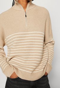 Beżowy sweter z zamkiem na szyi, z pionowym prążkowanym wzorem i poziomymi białymi paskami, o luźnym kroju i złożonymi mankietami.