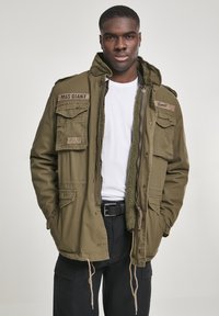 Olive green M65 jacket with a hood, multiple pockets, fleece lining, and patches. Features a zipper and adjustable drawstring at the hem.