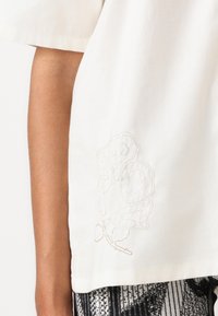 Top in cotone bianco con maniche corte, caratterizzato da un delicato ricamo floreale beige sul lato sinistro e un orlo dritto.