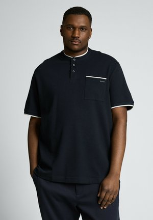 Mężczyzna o krótkich włosach, ubrany w czarną koszulę henley z krótkim rękawem z białym obszyciem i kieszenią na piersi, stoi na tle neutralnego, szarego tła.