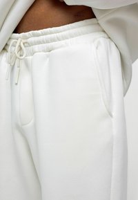 Witte joggingsbroek met een elastische tailleband en trekkoord. Beschikt over zijzakken en een zachte, gladde textuur. Close-up weergave.