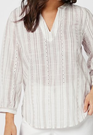 Femme portant un chemisier rayé blanc et rose clair avec des détails en œillets et une encolure fendue, associé à un pantalon blanc.