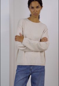 Beige pullovertröja med långa ärmar, rund halsringning, mjuk textur, avslappnad passform, tillsammans med blå jeans, stående framför en neutral bakgrund.