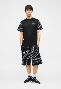 Άνδρας που στέκεται με τα χέρια στις τσέπες, φοράει μαύρο t-shirt Versace Jeans Couture, ταιριαστά σορτς με λευκό κείμενο, λευκές κάλτσες και μαύρα αθλητικά παπούτσια.