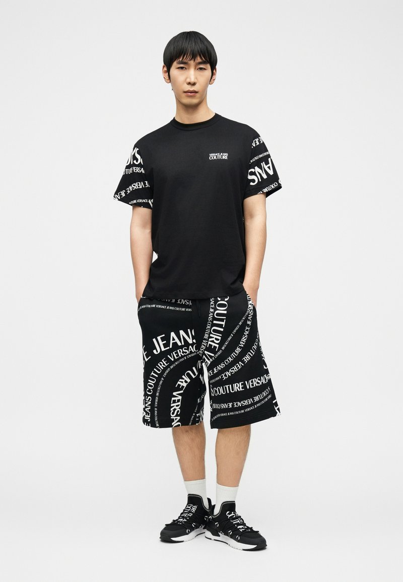 Άνδρας που στέκεται με τα χέρια στις τσέπες, φοράει μαύρο t-shirt Versace Jeans Couture, ταιριαστά σορτς με λευκό κείμενο, λευκές κάλτσες και μαύρα αθλητικά παπούτσια.