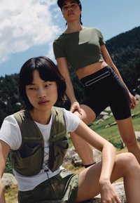 Two women outdoors in sporty clothing, one sitting on a rock wearing a white shirt and vest, the other standing in a green crop top and black shorts.