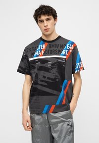 Νεαρός άνδρας που φοράει μαύρο graphic T-shirt BMW Motorsport με κόκκινες και μπλε ρίγες και γκρι παντελόνι Puma, στέκεται με το ένα χέρι στην τσέπη.