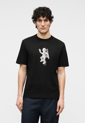 Uomo con capelli ricci che indossa una maglietta nera con la grafica di un astronauta in una tuta spaziale bianca, in piedi davanti a uno sfondo semplice.