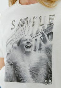 Witte T-shirt met korte mouwen met een zwart-witte afbeelding van een lachende vrouw, versierd met strass-steentjes die het woord "SMILE" spellen. Zachte textuur.