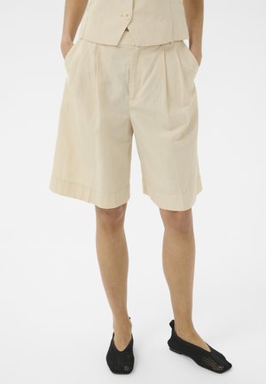 Licht beige knielange geplooide shorts gedragen met een bijpassend hesje en zwarte opengewerkte instapschoenen, persoon staand met handen in de zakken.