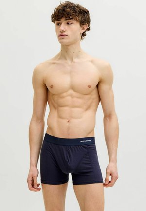 Jeune homme aux cheveux bouclés, torse nu, portant un boxer Jack & Jones bleu marine foncé sur un fond clair uni.
