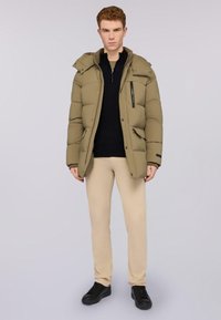 Giacca khaki imbottita con zip frontale, cappuccio rimovibile e due tasche frontali, indossata sopra una maglia nera e pantaloni beige. Sneakers nere.