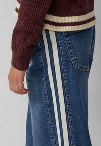 Maglione a maglia bordeaux con strisce crema all'orlo e ai polsini. Pantaloni in denim blu con due strisce verticali crema lungo i lati.