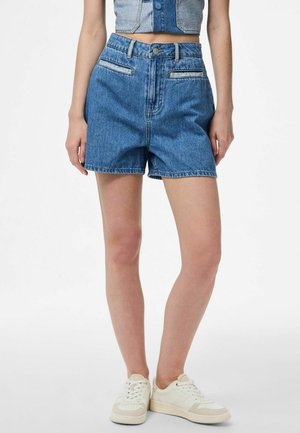 Kvinne iført blå denimhøytlivsshorts, kort denimvest og hvite joggesko, stående mot en ensfarget bakgrunn.