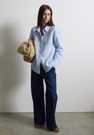 Vrouw draagt een lichtblauw shirt en brede, donkerblauwe jeans, houdt een geweven beige tas vast en staat tegen een effen witte muur.