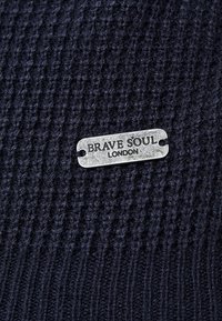 Marinblått stickat tyg med ett texturerat mönster och en silvermetalltagg som visar "BRAVE SOUL LONDON" fäst i mitten.