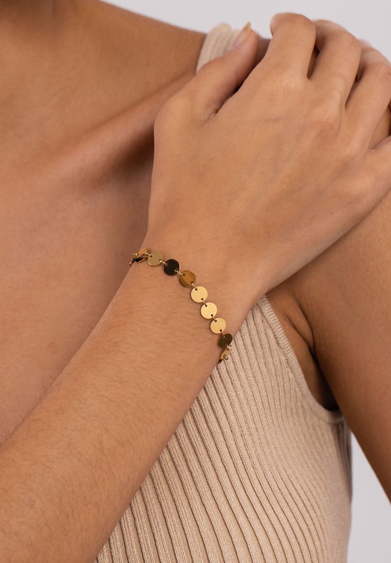 Poignet portant un bracelet à breloques en forme de disque en or sur peau nue, avec un haut beige côtelé sans manches visible sur le bras.