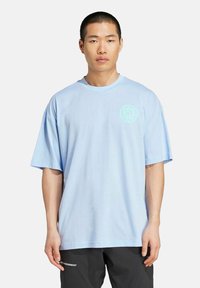 Camiseta de algodón azul claro con cuello redondo y mangas cortas, que presenta un sutil diseño gráfico en el lado izquierdo del pecho.