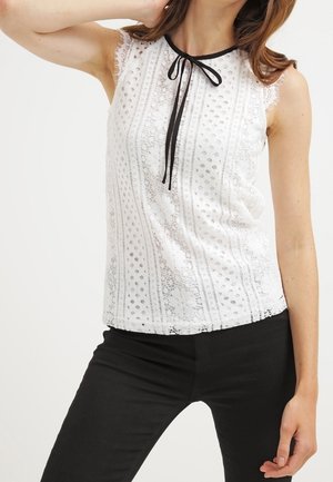 Nainen pukeutuneena hihattomaan valkoiseen pitsipaitaan, jossa on musta solmio kaulassa, yhdistettynä mustiin housuihin, poseeraa siten, että yksi käsi on kohotettuna ja lähellä päätä.