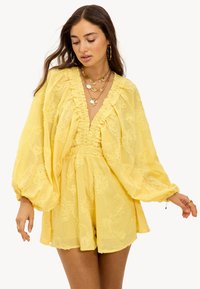 Combinaison jaune légère avec broderies florales, décolleté profond et manches bouffantes. Le tissu est semi-transparent avec une finition texturée.
