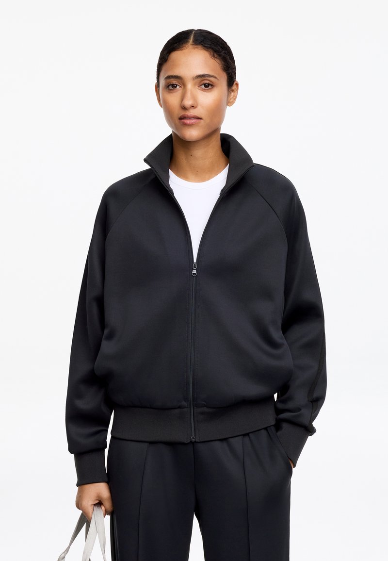 Femme aux cheveux foncés portant une veste noire zippée sur une chemise blanche, tenant un sac cabas gris clair sur un fond blanc uni.